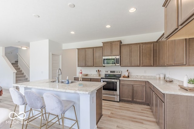 kitchen featuring stainless steel appliances, sink, a kitchen island with sink, light stone countertops, and light hardwood / wood-style flooring