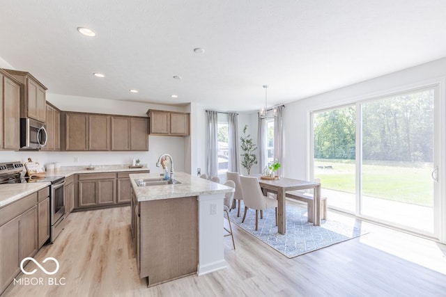 kitchen with sink, an island with sink, light wood-type flooring, appliances with stainless steel finishes, and decorative light fixtures