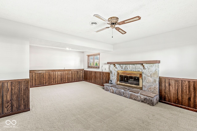 unfurnished living room featuring wood walls, light carpet, a stone fireplace, ceiling fan, and a textured ceiling