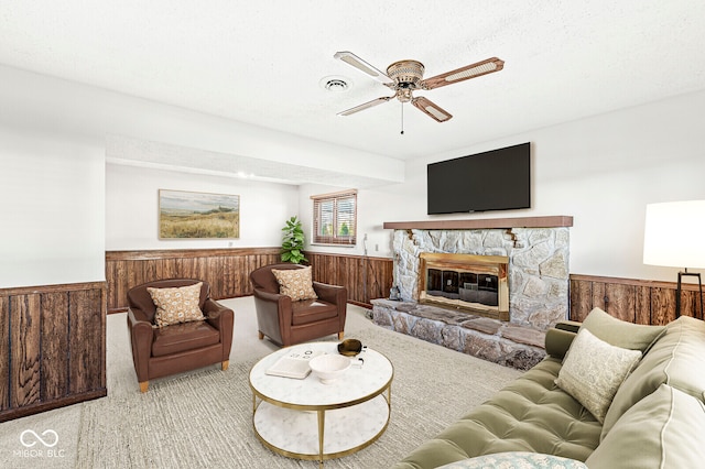 living room featuring ceiling fan, a stone fireplace, wood walls, light colored carpet, and a textured ceiling