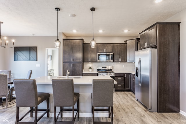 kitchen with dark brown cabinetry, stainless steel appliances, a kitchen island with sink, pendant lighting, and light hardwood / wood-style flooring