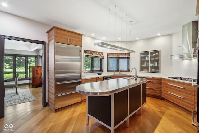 kitchen with light wood-type flooring, an island with sink, stainless steel appliances, and wall chimney range hood