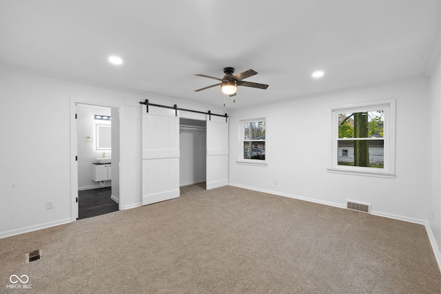 unfurnished bedroom featuring ensuite bath, ceiling fan, a barn door, carpet floors, and a closet