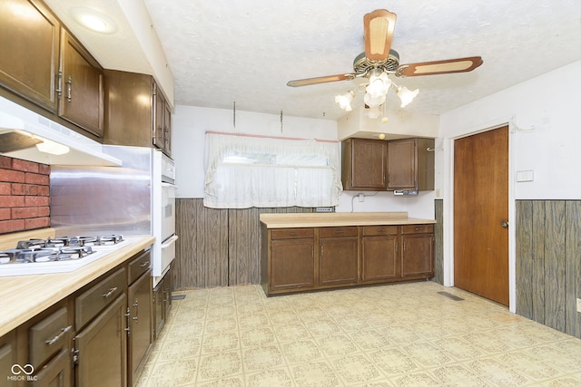 kitchen featuring a textured ceiling, white oven, ceiling fan, stainless steel gas stovetop, and wood walls