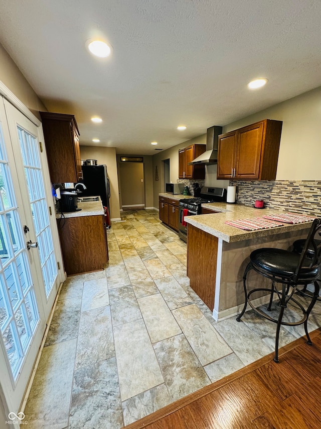 kitchen with kitchen peninsula, french doors, wall chimney range hood, a breakfast bar area, and stainless steel range with gas stovetop