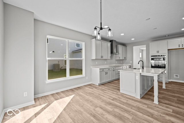 kitchen with pendant lighting, light hardwood / wood-style flooring, a kitchen island with sink, and wall chimney exhaust hood