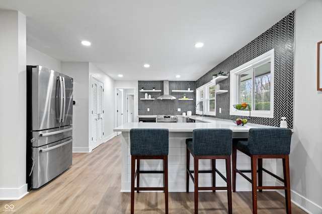 kitchen featuring kitchen peninsula, stainless steel appliances, wall chimney range hood, light hardwood / wood-style flooring, and a breakfast bar area