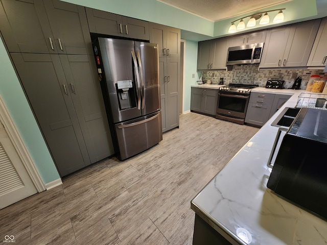 kitchen with a textured ceiling, gray cabinets, stainless steel appliances, light stone countertops, and backsplash