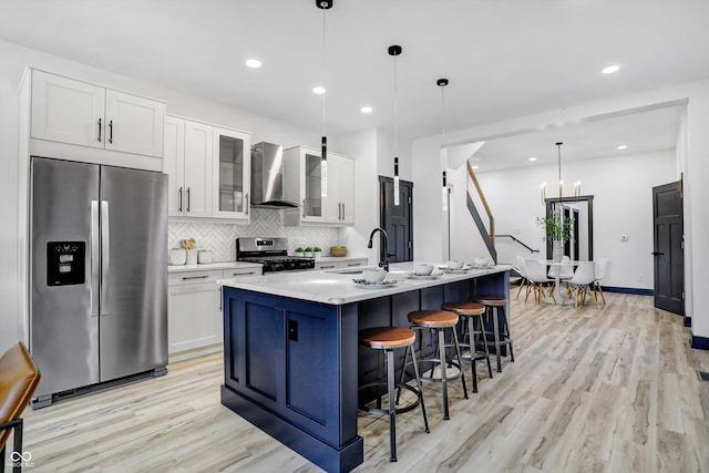 kitchen featuring stainless steel appliances, wall chimney range hood, pendant lighting, a center island with sink, and light wood-type flooring