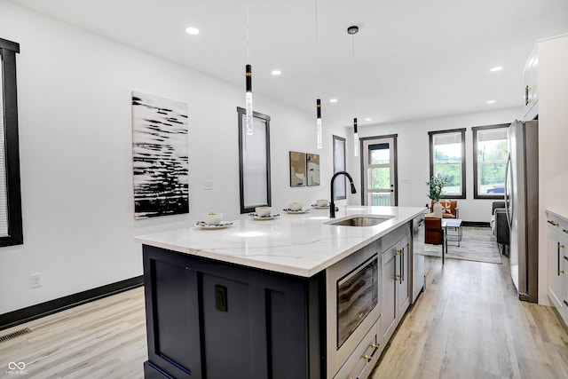 kitchen featuring hanging light fixtures, light stone counters, light hardwood / wood-style flooring, stainless steel fridge, and a kitchen island with sink