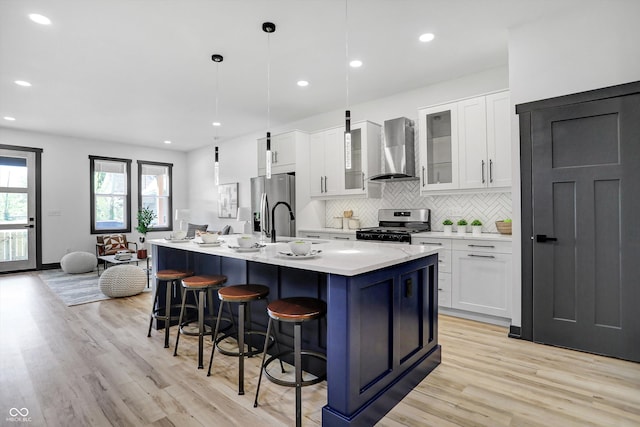 kitchen with appliances with stainless steel finishes, light wood-type flooring, wall chimney exhaust hood, a kitchen island with sink, and decorative light fixtures