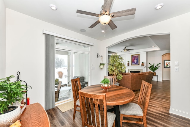 dining area with a tiled fireplace, ceiling fan, and dark wood-type flooring