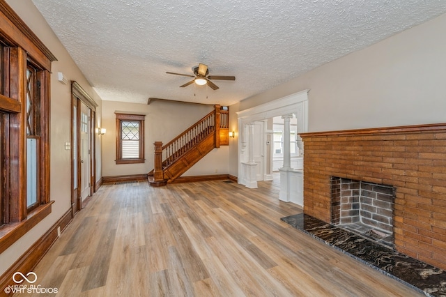 unfurnished living room featuring a brick fireplace, light hardwood / wood-style flooring, ceiling fan, a textured ceiling, and decorative columns