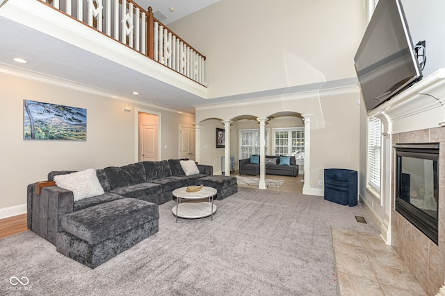 living room featuring a towering ceiling, ornate columns, light colored carpet, crown molding, and a tiled fireplace