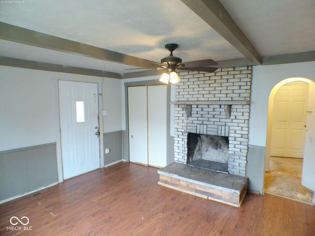 unfurnished living room featuring beam ceiling, ceiling fan, wood-type flooring, and a brick fireplace