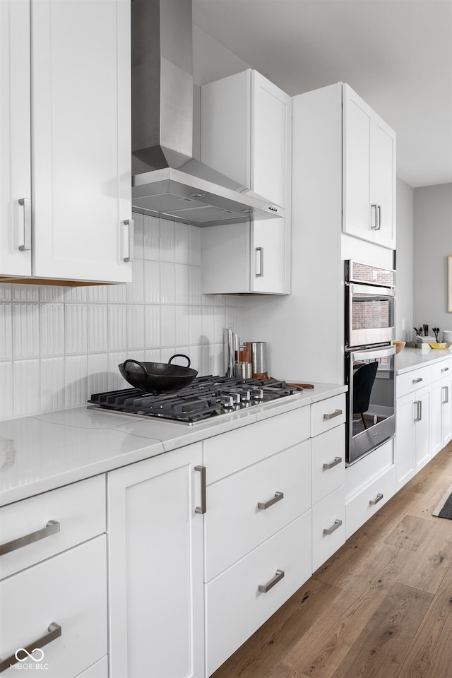 kitchen featuring light stone countertops, appliances with stainless steel finishes, wall chimney exhaust hood, light hardwood / wood-style floors, and white cabinetry