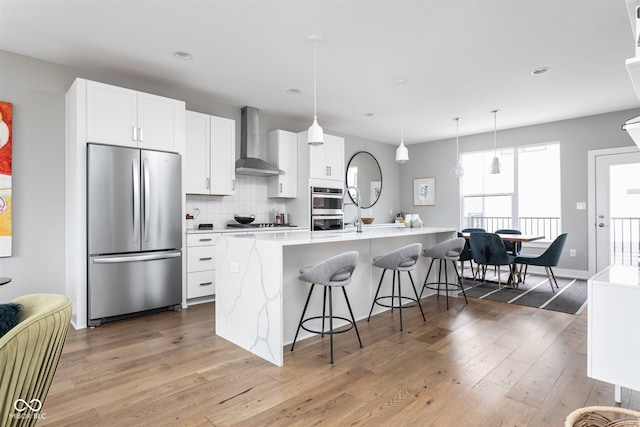 kitchen with wall chimney range hood, appliances with stainless steel finishes, a kitchen island with sink, white cabinets, and hardwood / wood-style flooring