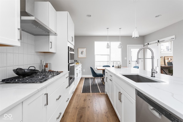 kitchen with wall chimney range hood, sink, a barn door, appliances with stainless steel finishes, and white cabinetry