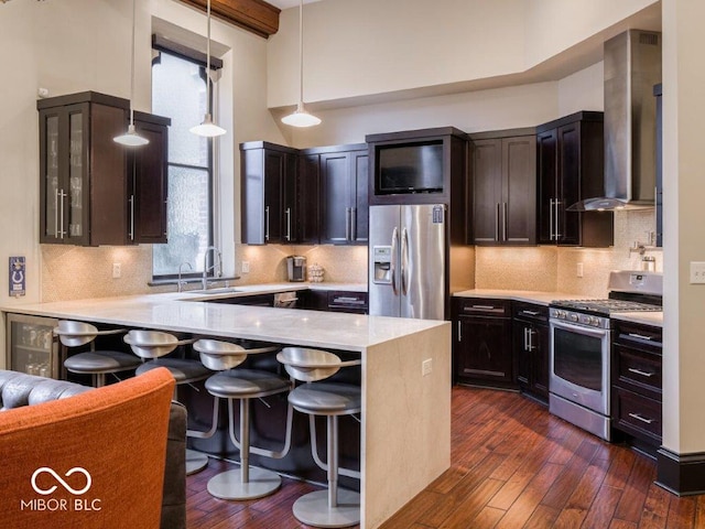 kitchen featuring pendant lighting, a breakfast bar, wall chimney range hood, appliances with stainless steel finishes, and dark hardwood / wood-style flooring