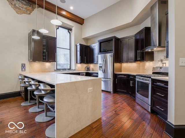 kitchen with dark wood-type flooring, a kitchen breakfast bar, wall chimney exhaust hood, appliances with stainless steel finishes, and kitchen peninsula