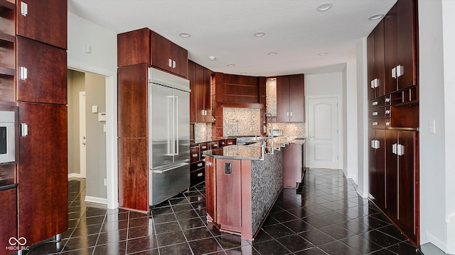 kitchen with backsplash, light stone counters, dark tile patterned floors, a center island, and built in fridge