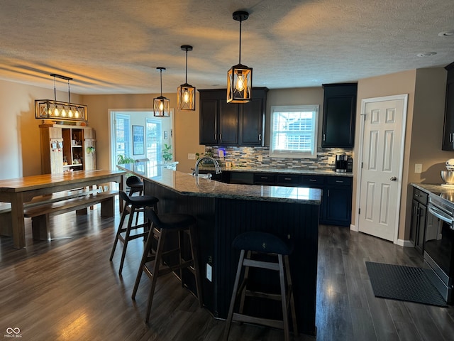 kitchen featuring a kitchen bar, decorative backsplash, a kitchen island with sink, dark stone countertops, and hanging light fixtures