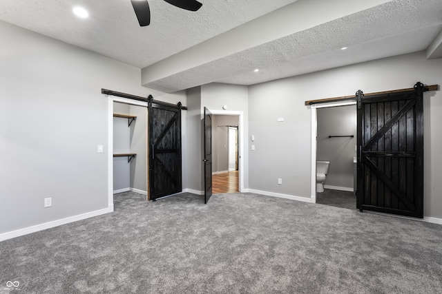 unfurnished bedroom featuring dark carpet, ensuite bathroom, ceiling fan, a barn door, and a textured ceiling