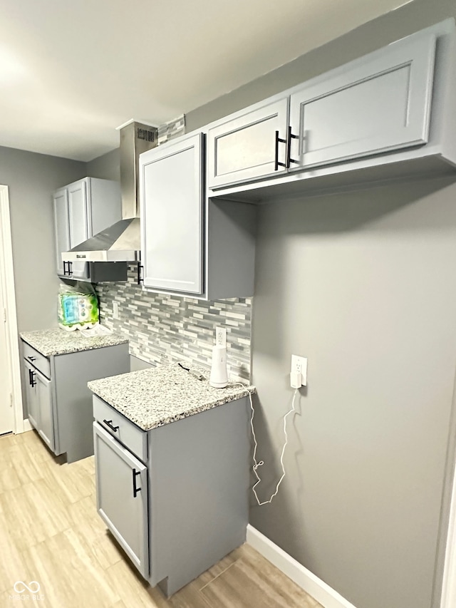 kitchen with light stone countertops, light wood-type flooring, tasteful backsplash, wall chimney range hood, and gray cabinets