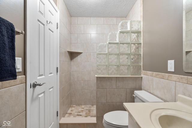 bathroom with a textured ceiling, toilet, tiled shower, vanity, and tile walls