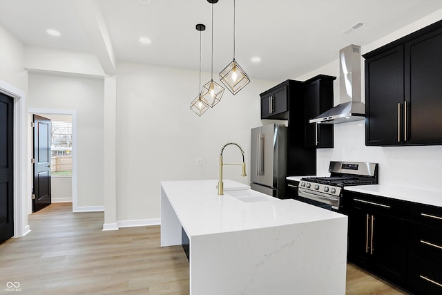 kitchen with stainless steel appliances, a kitchen island with sink, wall chimney range hood, pendant lighting, and light hardwood / wood-style flooring