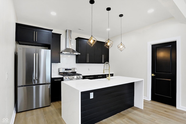 kitchen featuring a kitchen island with sink, wall chimney exhaust hood, light wood-type flooring, appliances with stainless steel finishes, and decorative light fixtures