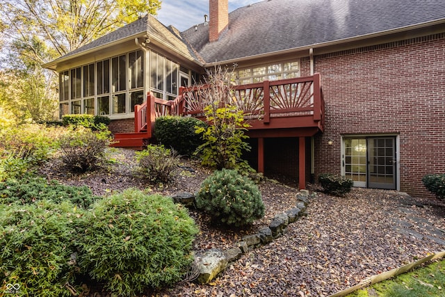 rear view of property featuring a sunroom, a shingled roof, a wooden deck, brick siding, and a chimney