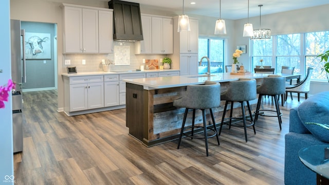 kitchen with a center island with sink, white cabinets, hanging light fixtures, and custom range hood