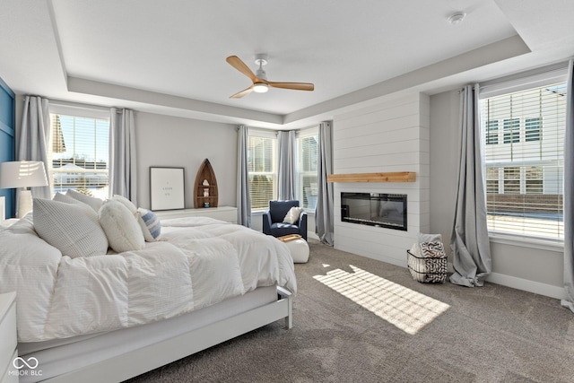 bedroom with carpet flooring, ceiling fan, a large fireplace, multiple windows, and a tray ceiling