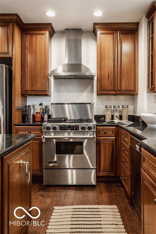 kitchen featuring dark stone countertops, dark wood-type flooring, stainless steel appliances, and wall chimney range hood