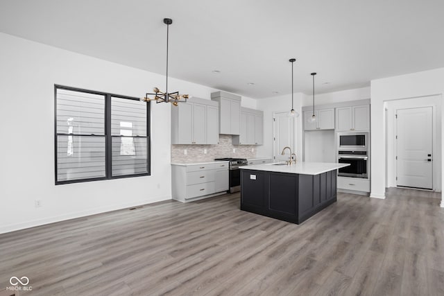 kitchen with appliances with stainless steel finishes, tasteful backsplash, a kitchen island with sink, decorative light fixtures, and light hardwood / wood-style flooring