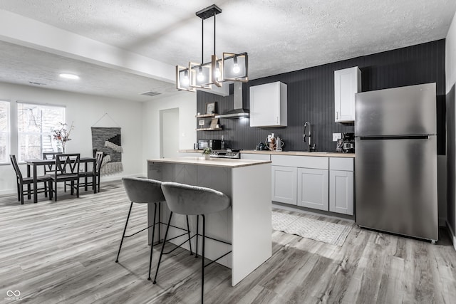 kitchen with stainless steel refrigerator, white cabinetry, wall chimney range hood, pendant lighting, and light hardwood / wood-style floors