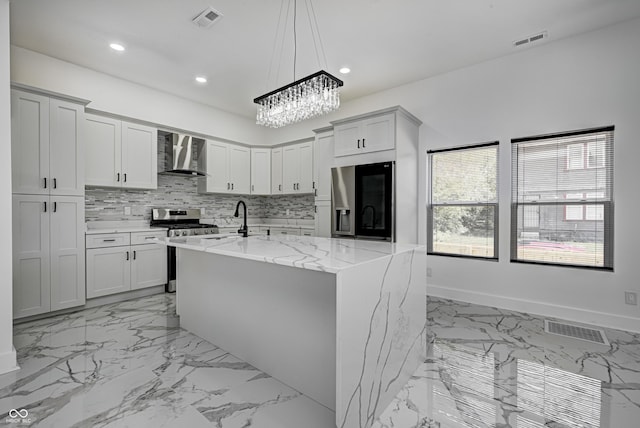 kitchen with stainless steel stove, wall chimney exhaust hood, an island with sink, refrigerator with ice dispenser, and light stone counters