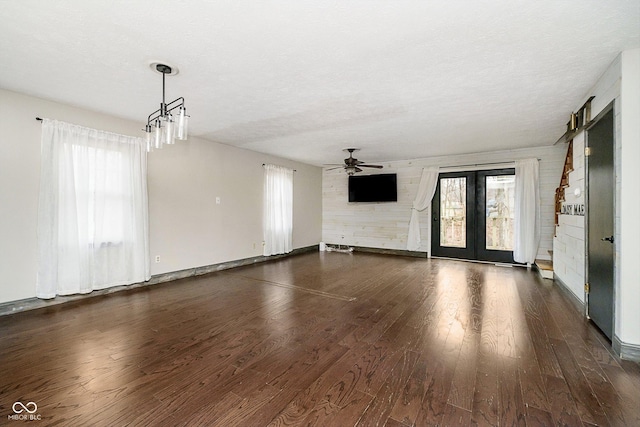 unfurnished living room featuring french doors, dark hardwood / wood-style floors, a textured ceiling, wooden walls, and ceiling fan with notable chandelier