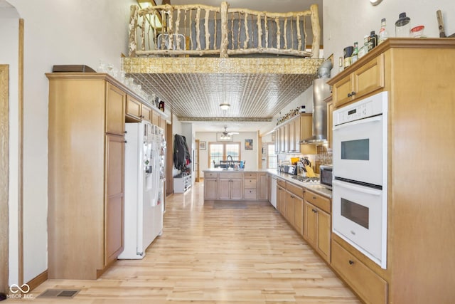 kitchen with white appliances, light wood-type flooring, kitchen peninsula, wall chimney exhaust hood, and light brown cabinets