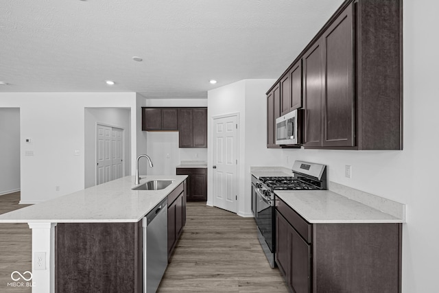 kitchen with appliances with stainless steel finishes, an island with sink, sink, dark brown cabinets, and light hardwood / wood-style flooring