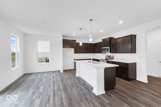 kitchen with decorative light fixtures, light countertops, appliances with stainless steel finishes, a kitchen island with sink, and a sink