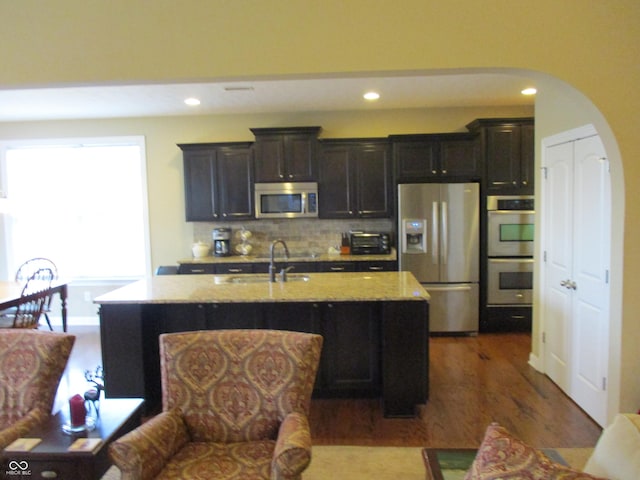 kitchen with light stone countertops, sink, hardwood / wood-style floors, an island with sink, and appliances with stainless steel finishes