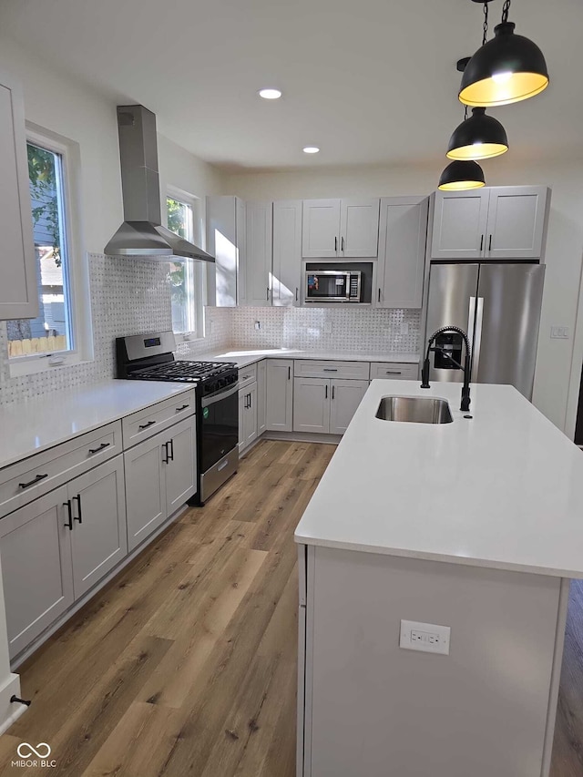 kitchen with sink, wall chimney exhaust hood, stainless steel appliances, pendant lighting, and a kitchen island with sink