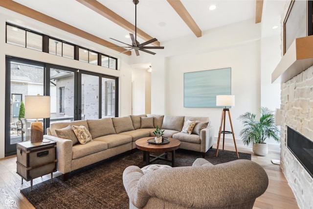 living room featuring beam ceiling, light wood-type flooring, ceiling fan, a fireplace, and a high ceiling