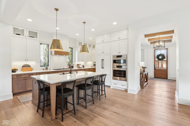 kitchen featuring white cabinetry, hanging light fixtures, a center island with sink, light wood-type flooring, and stainless steel appliances