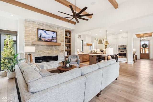 living room with beamed ceiling, ceiling fan, a stone fireplace, and light wood-type flooring