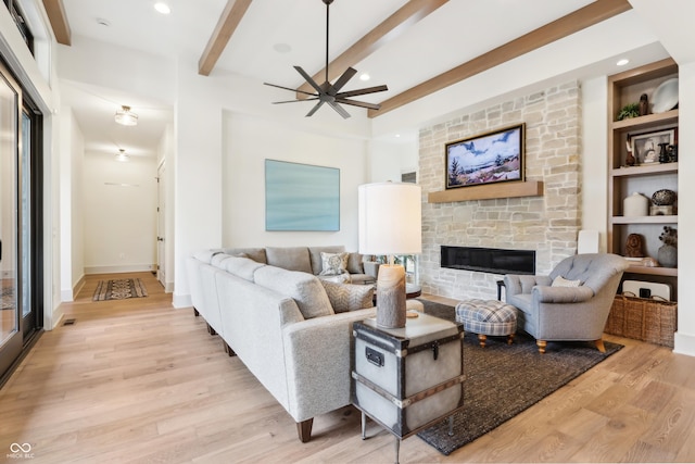 living room with a stone fireplace, built in features, beamed ceiling, ceiling fan, and light wood-type flooring