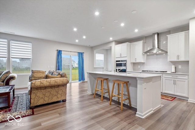 kitchen with double oven, wall chimney range hood, a center island with sink, white cabinets, and light hardwood / wood-style floors