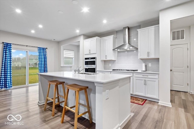 kitchen featuring white cabinetry, wall chimney exhaust hood, gas stovetop, a kitchen island with sink, and light wood-type flooring